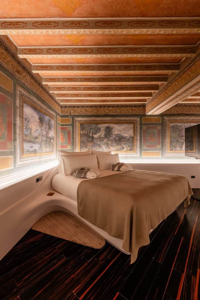 Hotel Romeo Roma bedroom with a painted timber beamed ceiling and a frieze of framed landscape frescoes above a flowing white bed base on striped ebony floorboards
