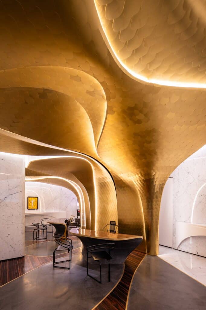 Golden scale-clad vaults flowing down into a column at the Hotel Romeo Roma reception, with an ebony desk, black chairs and white marble walls beyond