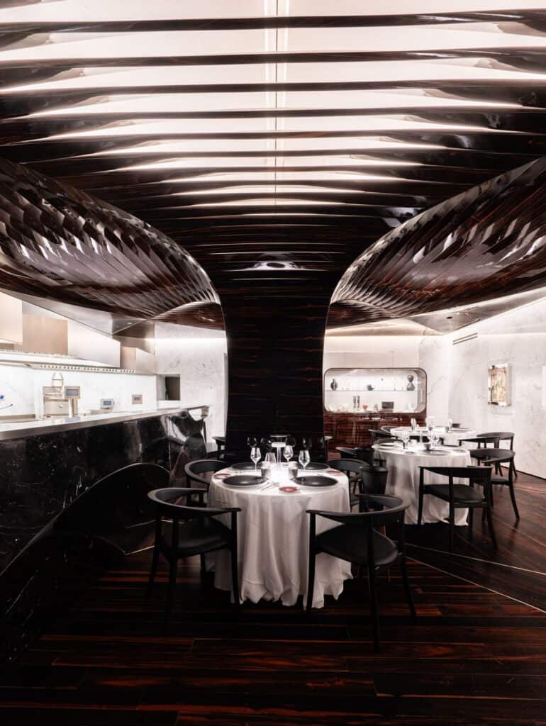 Il Ristorante Alain Ducasse at Hotel Romeo Roma, a dark column branching into ribbed reflective canopies above round tables laid with white cloths and a black marble counter