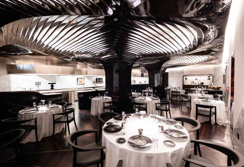 Wide view of the Alain Ducasse restaurant at Hotel Romeo Roma, silver tree-like ceiling ribs spreading from dark columns over white-clothed tables and the open kitchen behind