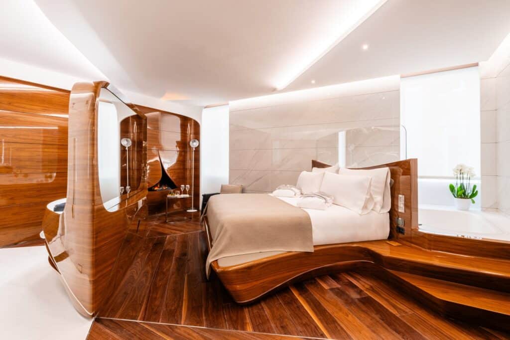 Deluxe room at Hotel Romeo Roma with a bed rising from a stepped honey-coloured wood platform, a curved wooden screen and a white marble bathroom behind glass