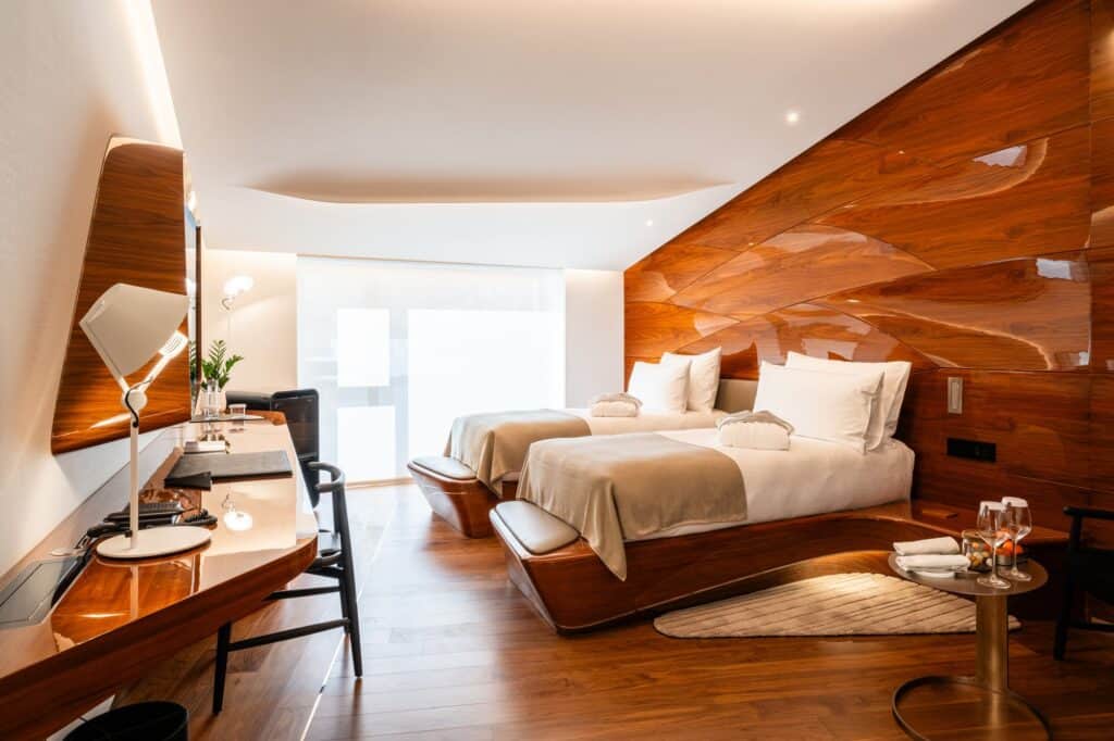 Twin room at Hotel Romeo Roma with two beds on curved wooden bases, a sculpted glossy wood wall, a built-in desk with a lamp and sheer curtains at the window