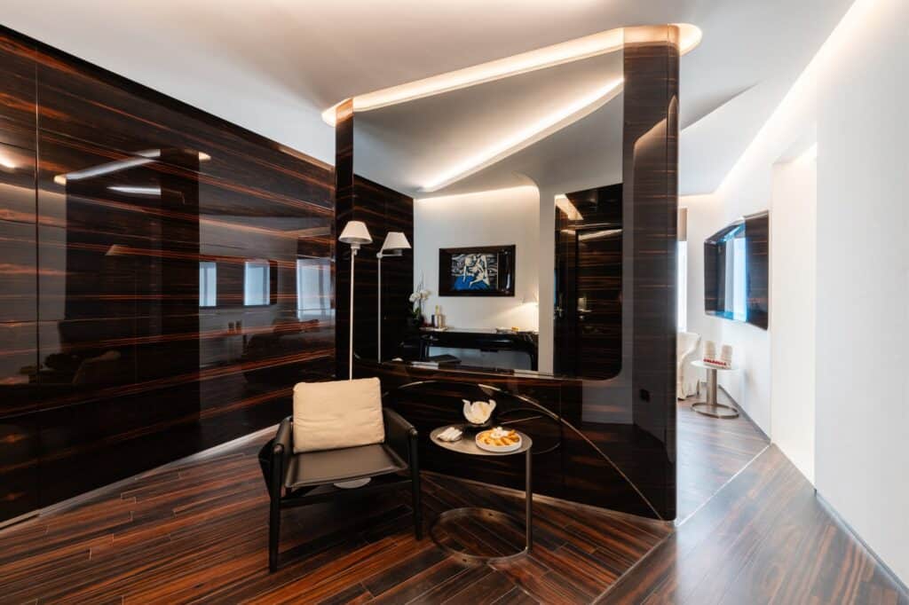 Sitting area of a Hotel Romeo Roma suite with glossy ebony wall panels, a black armchair and side table, lit ceiling coves and a white corridor beyond