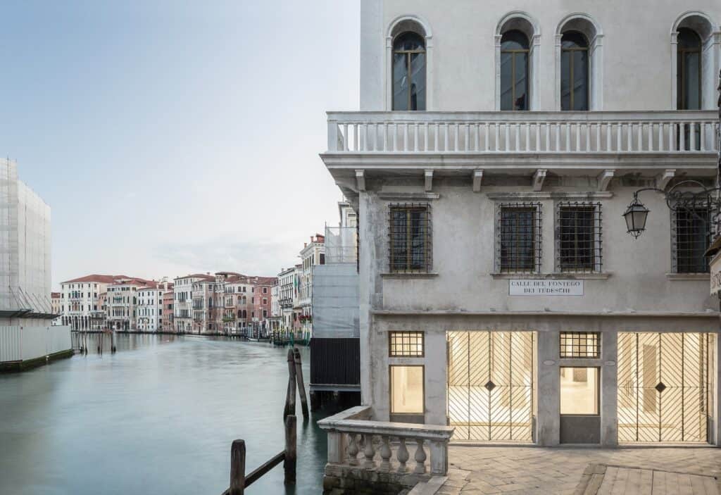 Corner of Il Fondaco dei Tedeschi on the Grand Canal, its pale facade with a stone balustrade balcony, arched windows and lit glazed shopfronts beside the water, palazzi lining the canal beyond