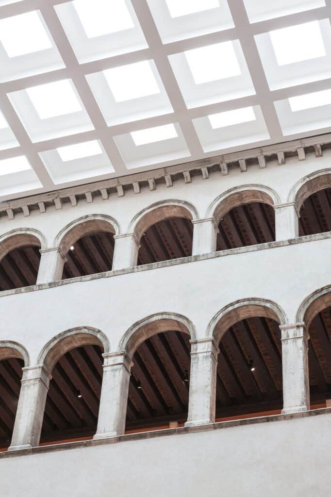Two tiers of round-arched stone arcades in the courtyard of Il Fondaco dei Tedeschi beneath a dentil cornice and the grid of the glazed skylight roof, dark timber ceilings visible behind the arches