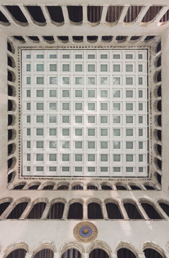 Looking straight up the courtyard of Il Fondaco dei Tedeschi to its square grid skylight, framed by tiers of stone arcades and a blue-and-gold clock face on the wall below