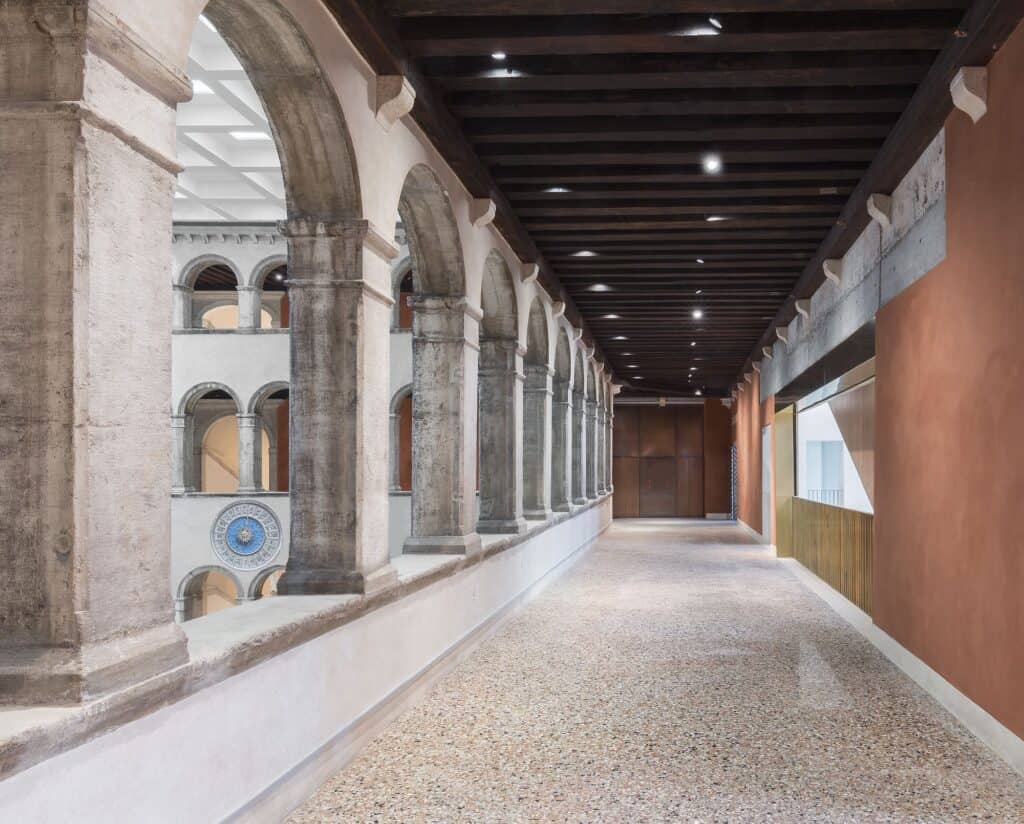 Upper loggia of Il Fondaco dei Tedeschi with weathered stone columns and arches open to the courtyard on the left, a speckled terrazzo floor, dark timber ceiling and terracotta-red wall on the right
