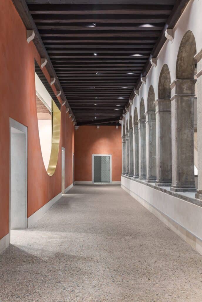 Long loggia corridor in Il Fondaco dei Tedeschi between a terracotta-red wall with a curved gold-lined cut-out and a row of stone columns, beneath a dark beamed ceiling