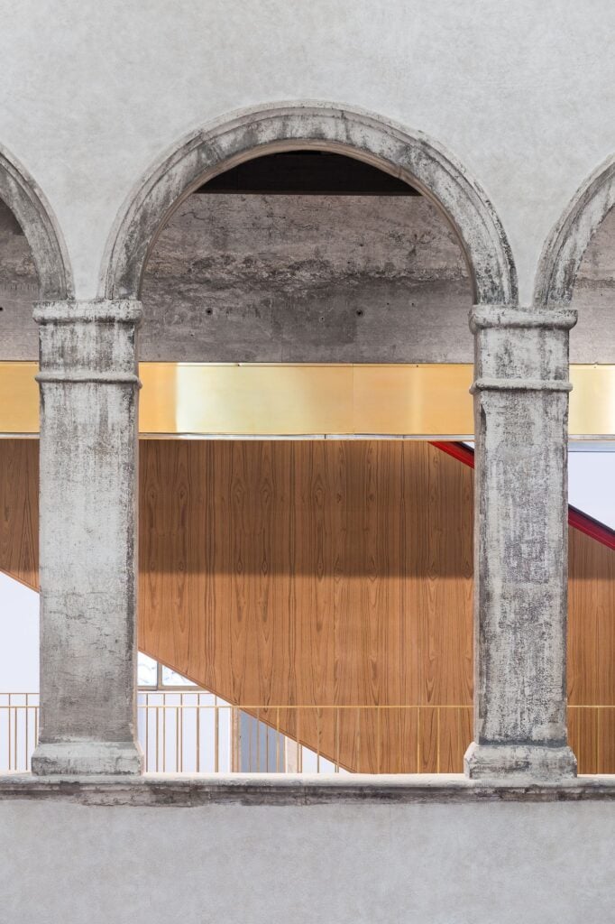 Pair of weathered stone columns and arches in Il Fondaco dei Tedeschi framing a gold band and the timber-panelled side of a new escalator rising behind