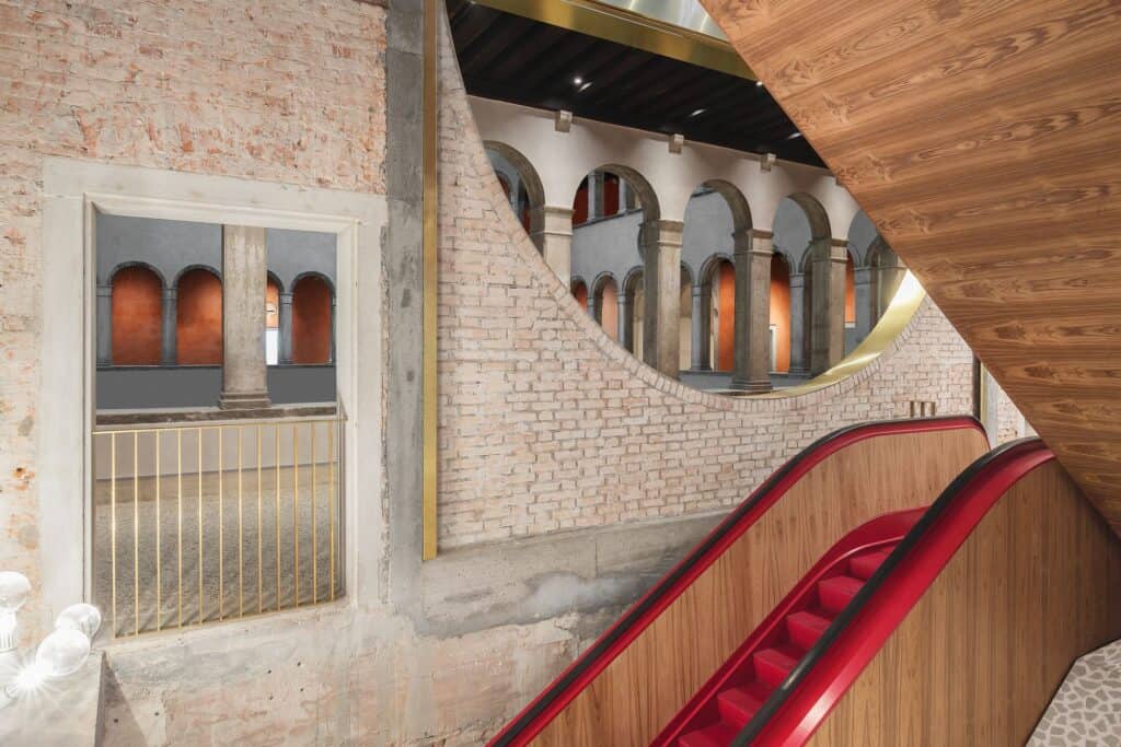 Red escalators with wood-panelled sides rising through Il Fondaco dei Tedeschi past exposed brick walls, a large circular opening revealing the courtyard arcades and a gold-railed window