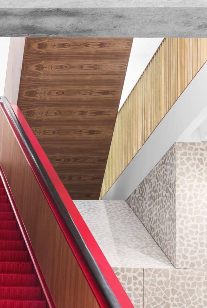 Bright red escalator steps and handrail beside a timber-panelled wall in Il Fondaco dei Tedeschi, angled timber soffits and a pale mosaic-like stone wall beyond