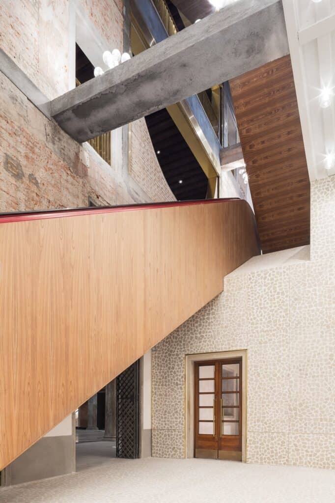 Wood-panelled escalator housing crossing a tall space in Il Fondaco dei Tedeschi beneath a bare concrete beam, exposed brick walls above and a glazed timber door in a pale stone wall below