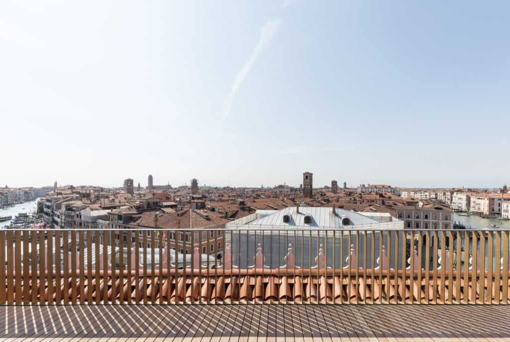Wooden roof terrace of Il Fondaco dei Tedeschi with a bronze-toned slatted balustrade looking over the terracotta rooftops and bell towers of Venice under a pale blue sky