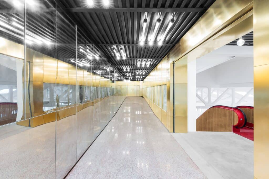Gold-clad corridor in Il Fondaco dei Tedeschi with a mirrored wall reflecting rows of lights, a polished terrazzo floor and a red escalator glimpsed through an opening on the right