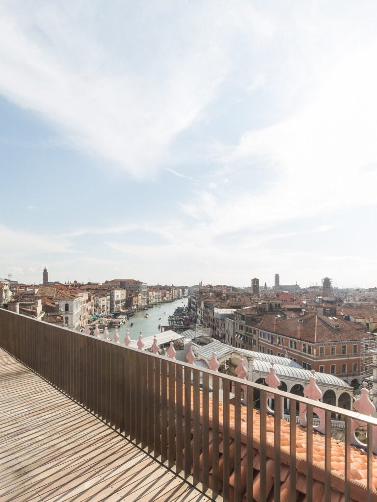 Timber deck and slatted balustrade of the Il Fondaco dei Tedeschi roof terrace, looking down the Grand Canal as it winds between palazzi and terracotta roofs under a wide cloudy sky