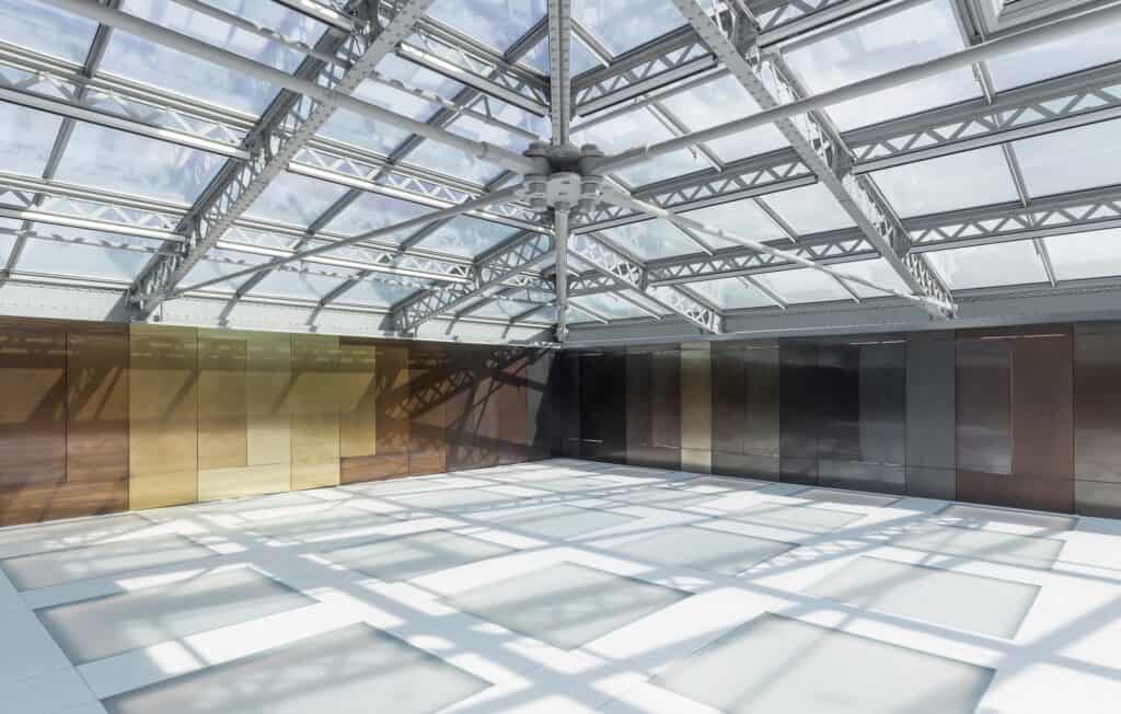 Sunlight casting the shadows of steel roof trusses across the translucent glass floor of the Il Fondaco dei Tedeschi event pavilion, metallic wall panels running around the room
