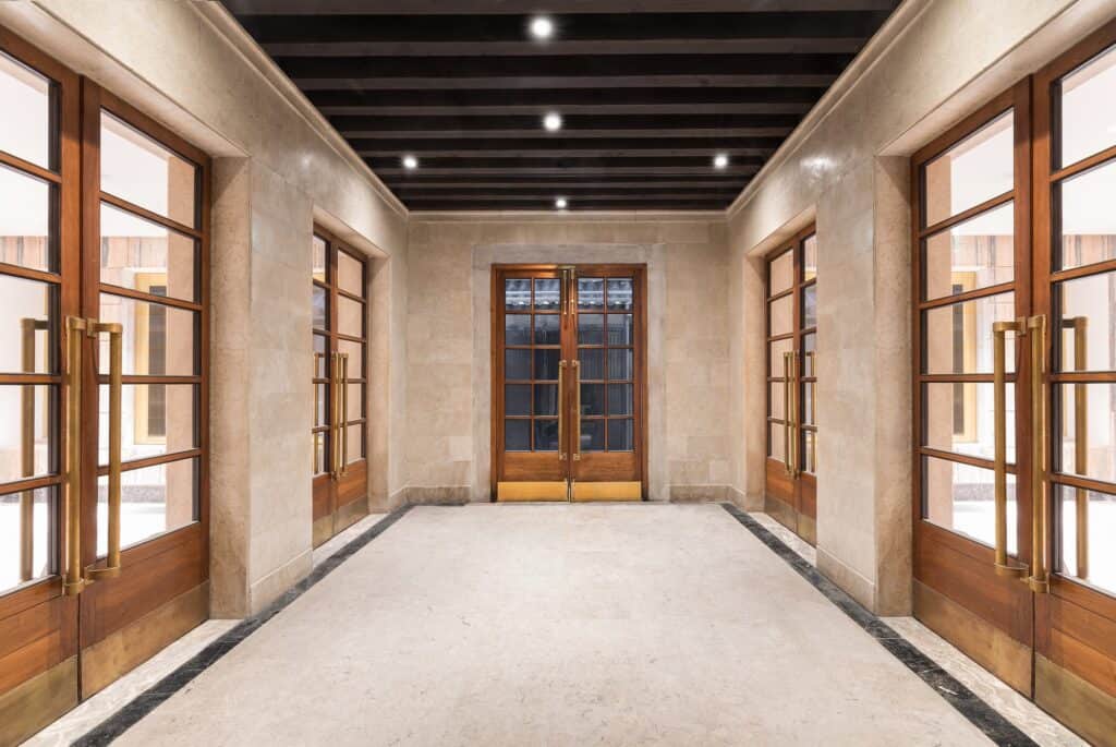 Pale stone corridor in Il Fondaco dei Tedeschi lined with tall timber-framed glazed doors with long brass handles, a terrazzo floor with dark borders and a dark beamed ceiling
