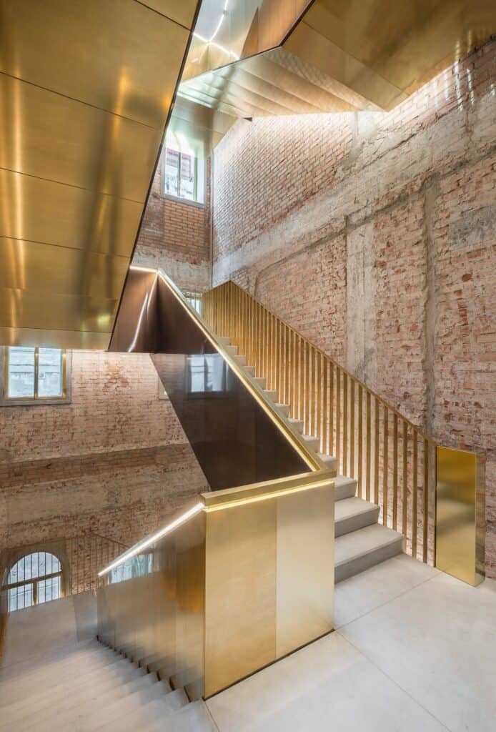 New staircase in Il Fondaco dei Tedeschi with a polished brass balustrade and gold-clad soffit rising between rough brick and concrete walls, lit by a high window