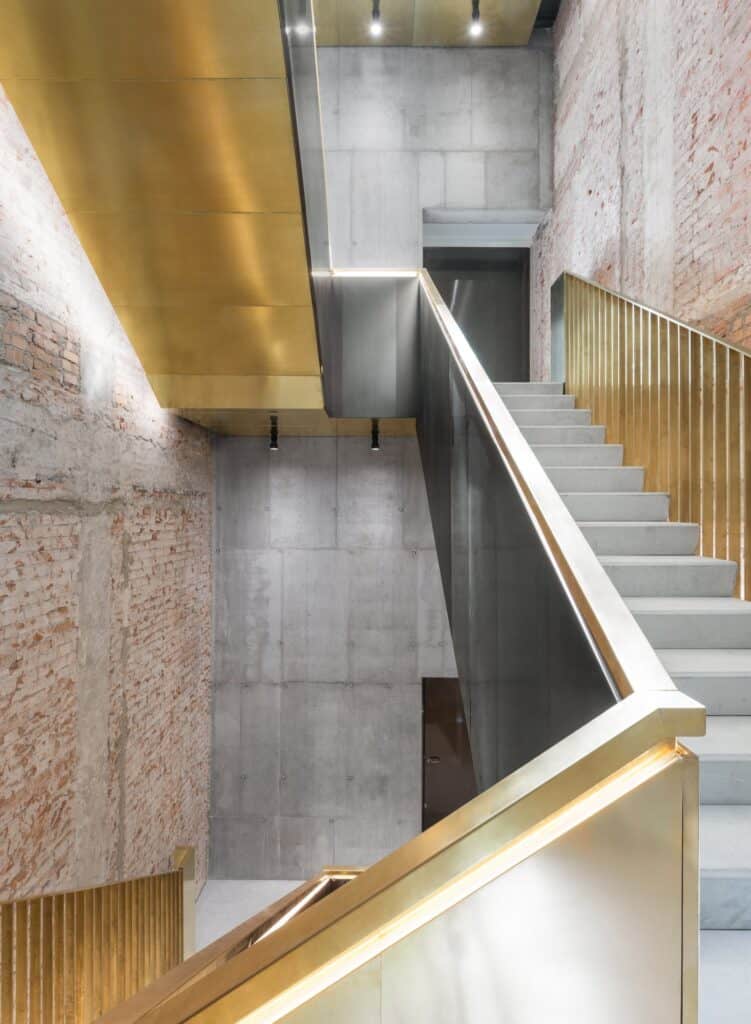 Concrete stair flights with brass balustrades and gold-lined soffits climbing through a shaft of exposed brick and board-marked concrete in Il Fondaco dei Tedeschi