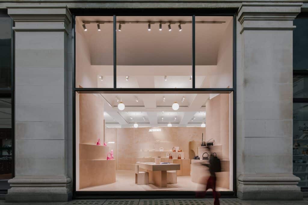 Evening view through the tall glazed shopfront of Jacquemus Store Selfridges London between stone pilasters, a passer-by blurred in front