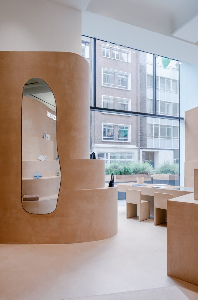 Rounded terracruda display tower with an irregular oval mirror reflecting the shelves, clay table and chairs at the window, Jacquemus Selfridges