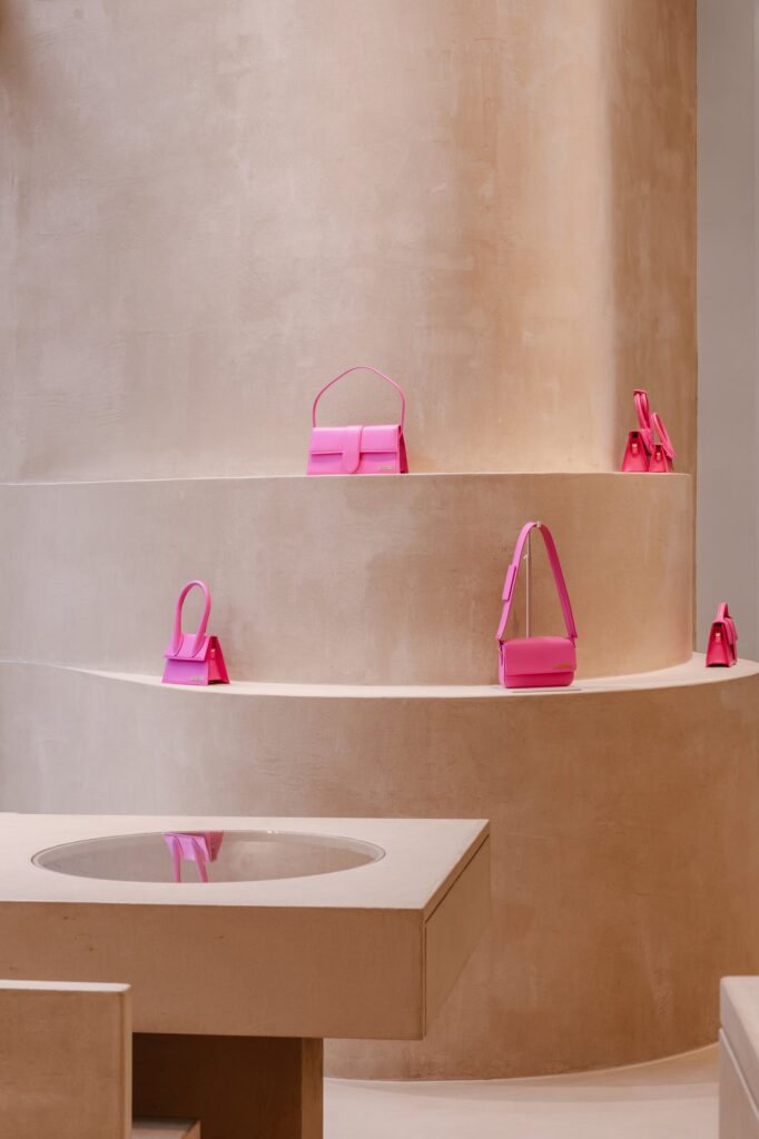 Bright pink Jacquemus bags on a curved terracruda ledge above a clay table with a round glass vitrine at Jacquemus Store Selfridges London