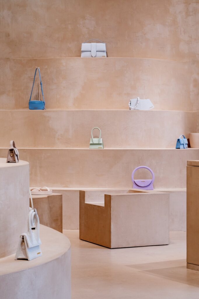 Wavy terracruda shelves rising in tiers with pastel Jacquemus bags, a lilac ring-handled bag on a chunky clay seat below, Selfridges London