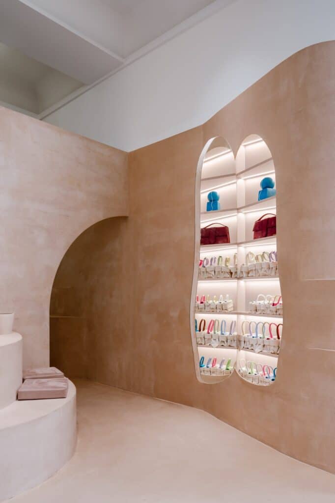 Two backlit pill-shaped niches stacked with colourful Jacquemus bags in a sweeping clay wall beside an arched passage at Selfridges London