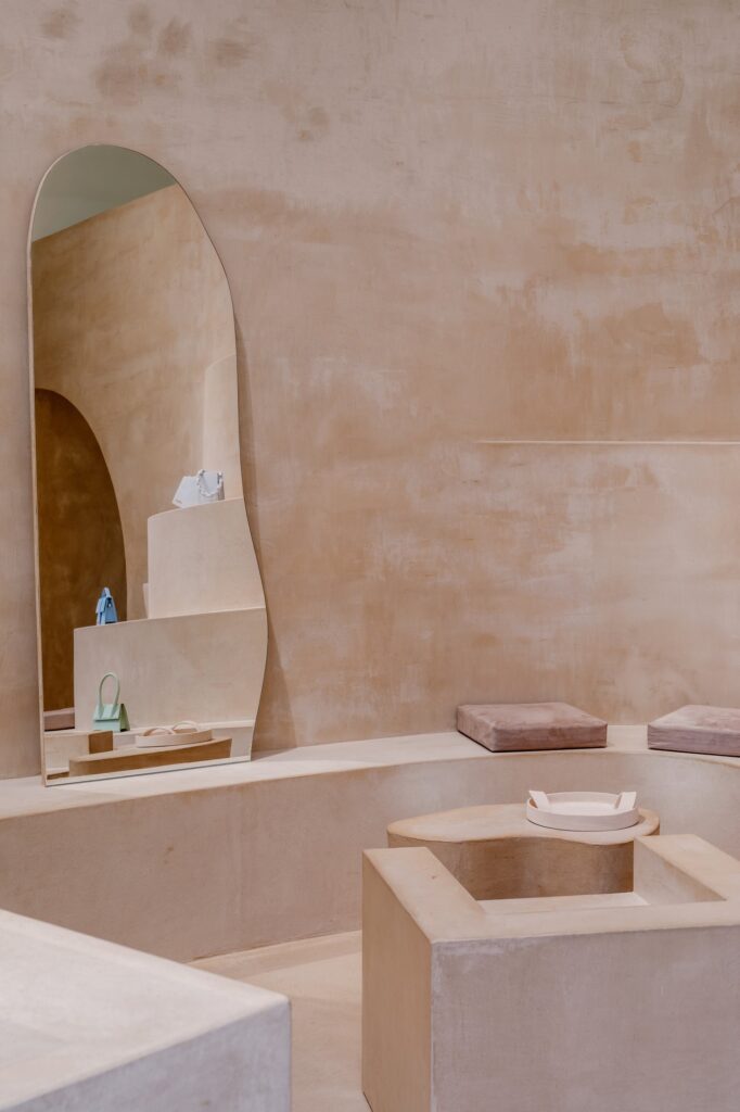 Tall irregular mirror leaning on a curved clay bench with dusty pink cushions, reflecting bags on stepped shelves at Jacquemus Selfridges