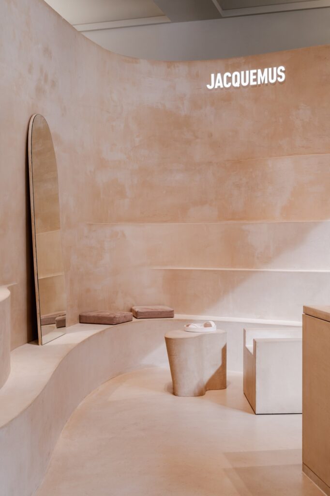 Curving clay bench with dusty pink cushions and an arched mirror beneath the illuminated Jacquemus sign, a sculpted clay side table in front