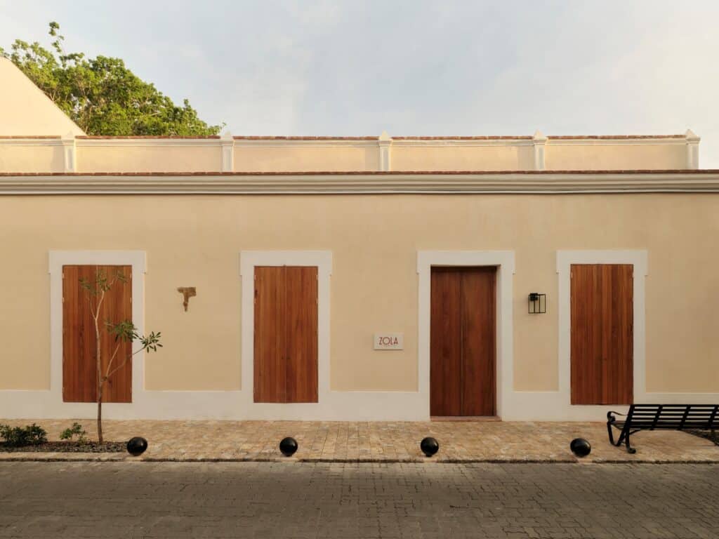 Restored single-storey colonial frontage of Kimpton Las Mercedes Hotel, a cream rendered wall with four stone-framed timber doors, black bollards and a bench on the pavement