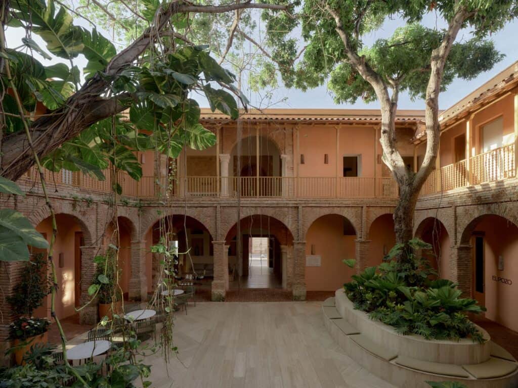 Central courtyard of Kimpton Las Mercedes Hotel, a mature tree rising over a brick-arched colonial arcade with a timber gallery above and planters on the stone paving