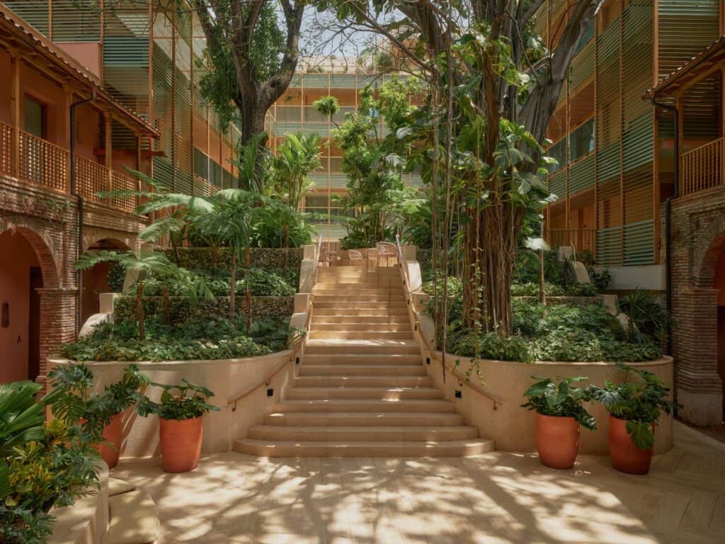 Wide stone staircase climbing between planted terraces in the raised courtyard of Kimpton Las Mercedes Hotel, framed by trees, terracotta pots and green-louvred galleries