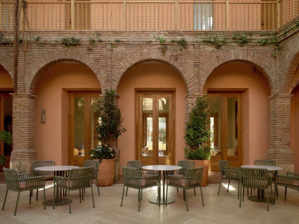 Brick arcade of the colonial courtyard at Kimpton Las Mercedes Hotel, three round arches with salmon-pink walls and tall timber doors behind café tables and green chairs