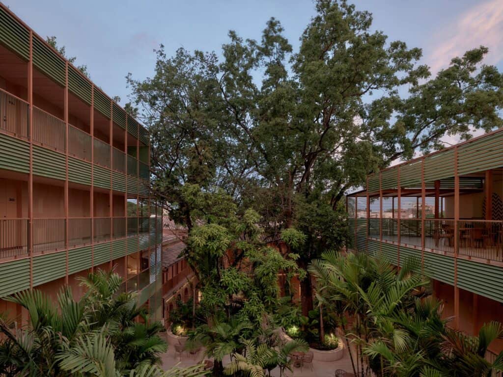 Courtyard of Kimpton Las Mercedes Hotel at dusk, a large tree and palms between new wings with open galleries screened by green horizontal louvres