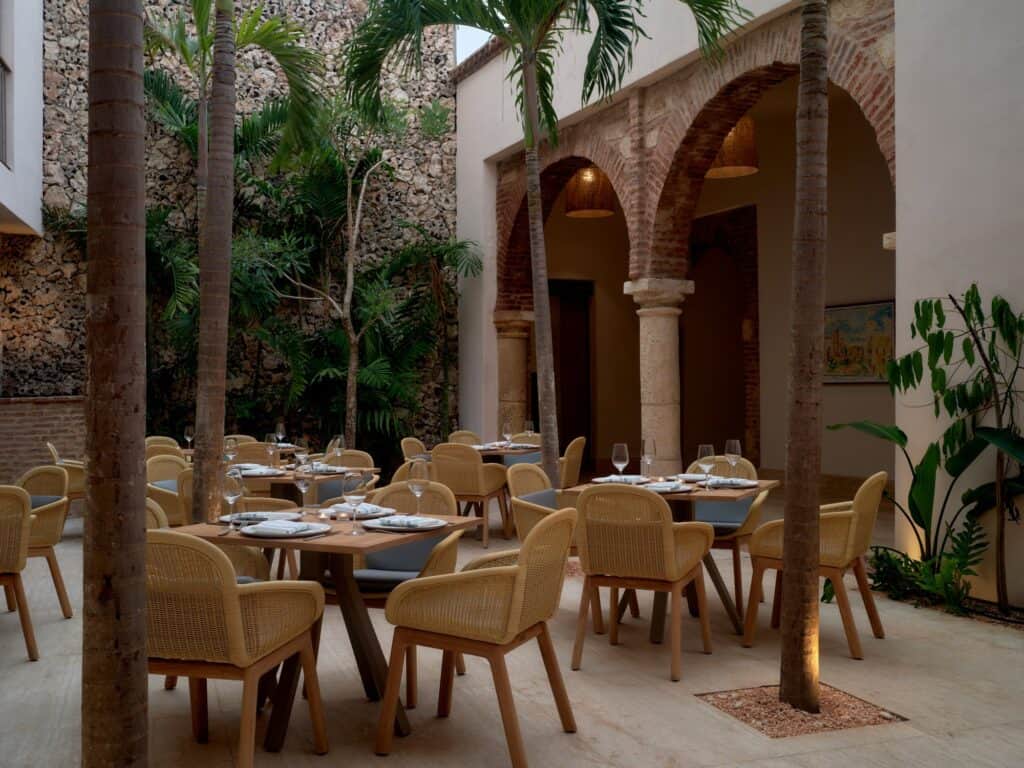 Restaurant in a courtyard at Kimpton Las Mercedes Hotel, rattan chairs and set tables between palm trunks beneath brick arches on stone columns and a rubble-stone wall