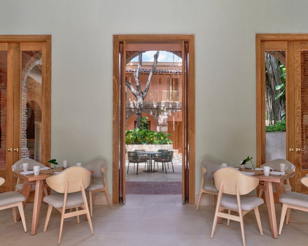 Dining room at Kimpton Las Mercedes Hotel with pale timber chairs and small tables between tall double doors framed in wood, one opening onto the arcaded courtyard