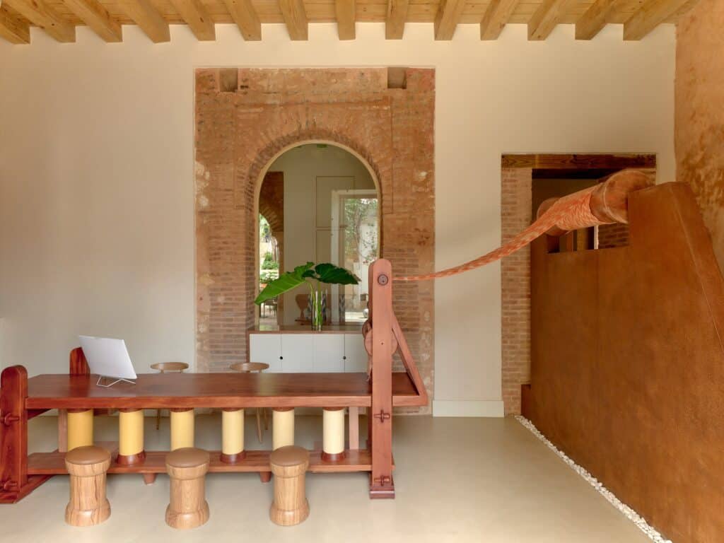 Lobby at Kimpton Las Mercedes Hotel under a timber-beamed ceiling, a loom-like timber desk with an orange woven textile stretched across to a roller on a rust-red wall, before an exposed brick arch