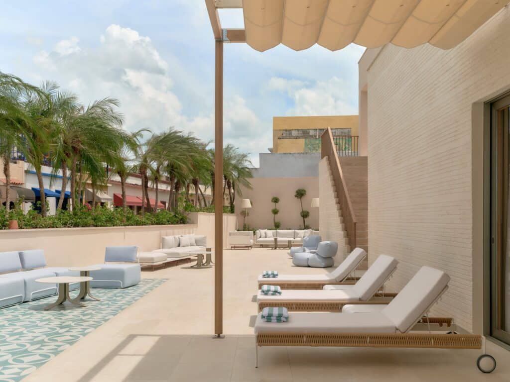 Rooftop terrace at Kimpton Las Mercedes Hotel with white sun loungers under a cream canvas pergola, blue patterned tiles and sofas, and palm trees above the parapet