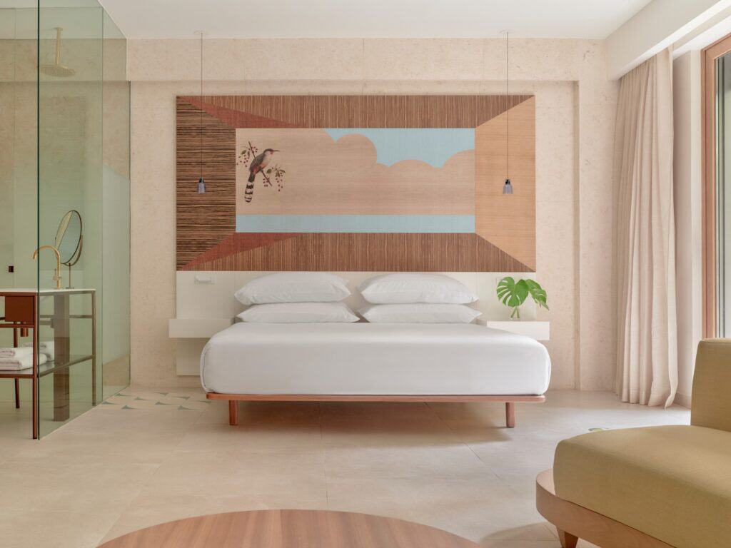 Guest room at Kimpton Las Mercedes Hotel with a white bed beneath a large marquetry panel of a bird and sky, travertine walls and a glass-walled shower to the side