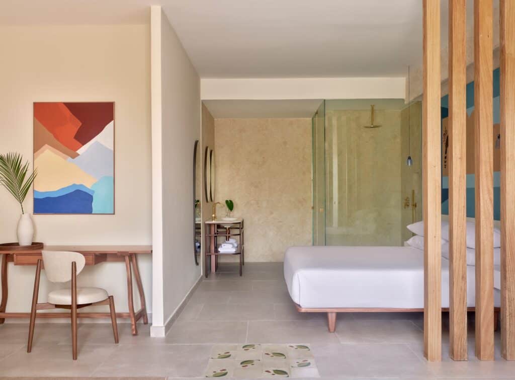 Guest room at Kimpton Las Mercedes Hotel with timber slat screen beside the bed, a writing desk under a colourful abstract painting and a travertine-lined bathroom beyond