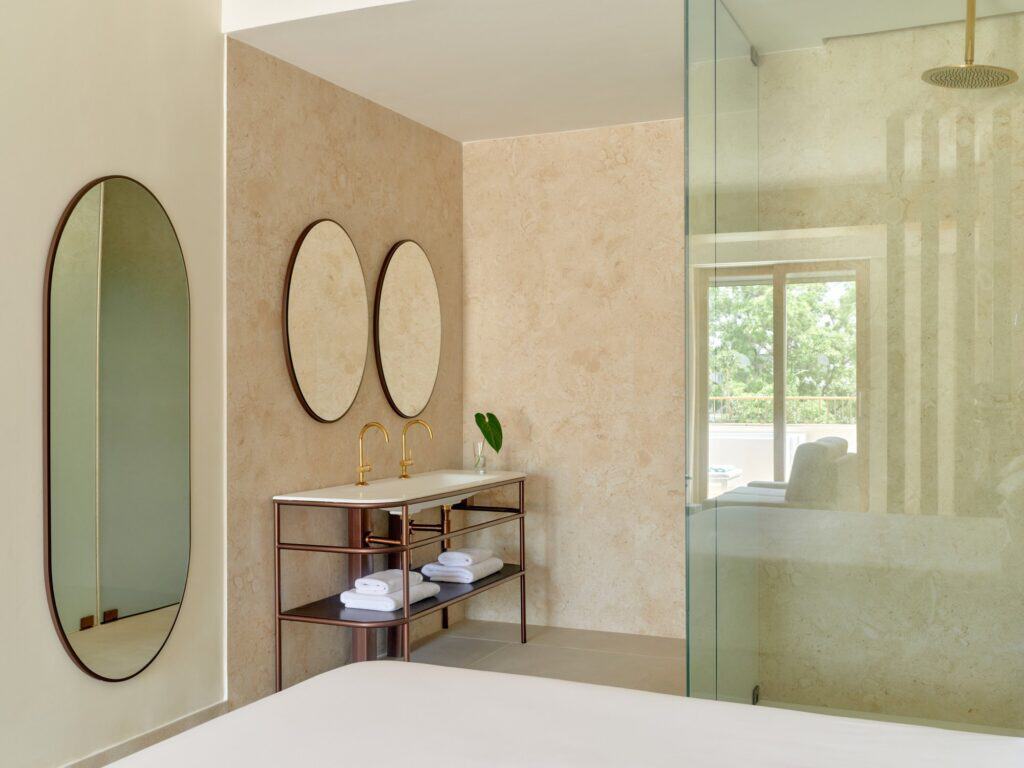 Bathroom at Kimpton Las Mercedes Hotel with travertine walls, a capsule-shaped mirror and two round mirrors over a slender metal washstand, beside a glass shower