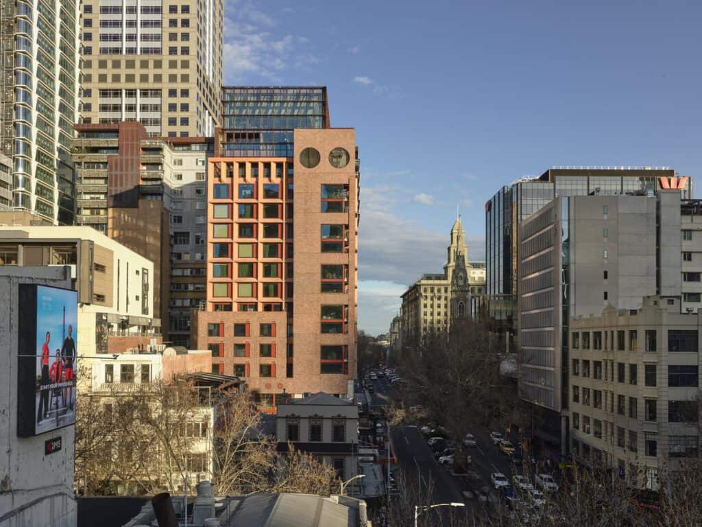 Melbourne Place Hotel rising in red brick and terracotta-tinted concrete above a tree-lined CBD street, two circular oculi near its top