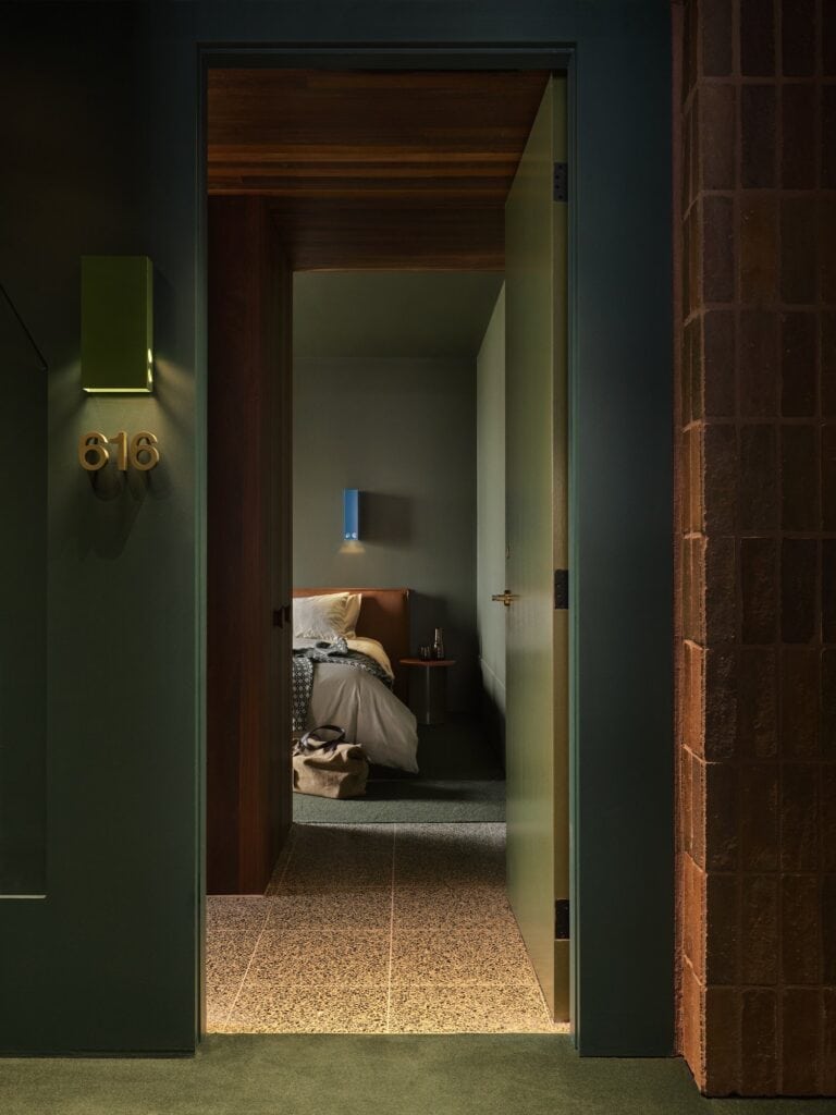 Green corridor outside room 616 at Melbourne Place Hotel, a brick column beside the open door and a terrazzo entry leading to the bed