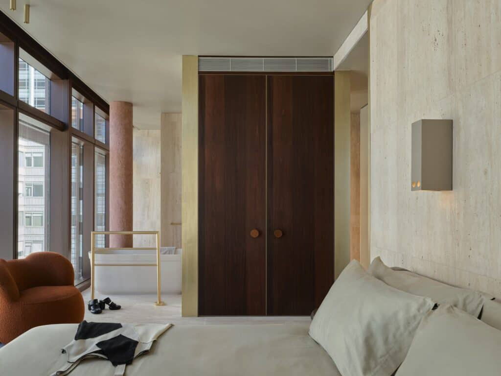 Penthouse bedroom at Melbourne Place Hotel with travertine walls, brass-framed jarrah wardrobe doors, a pink column and a freestanding bath