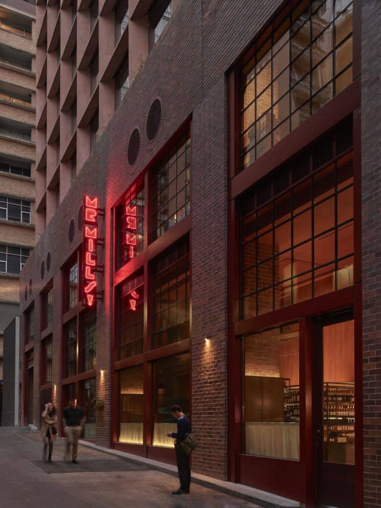 Dark red brick laneway frontage of Melbourne Place Hotel at dusk with tall steel-framed windows and the red neon Mr Mills sign