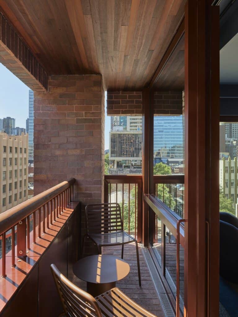 Brick-lined balcony at Melbourne Place Hotel with a timber soffit, red metal balustrade, two slatted chairs and a side table above the city