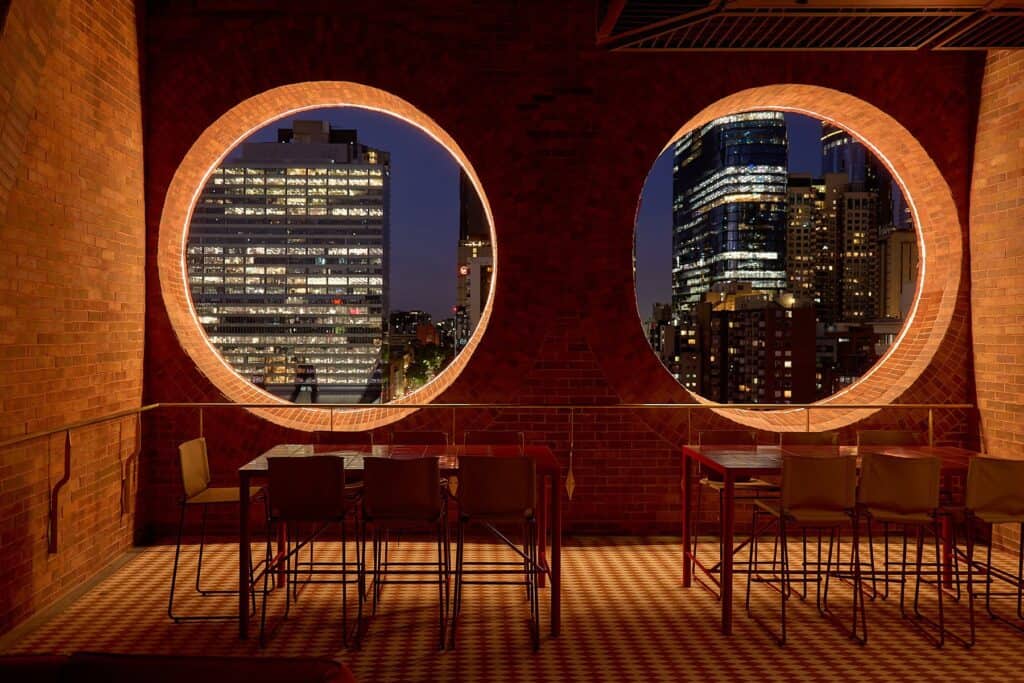 Twin glowing brick oculi at Mid Air, Melbourne Place Hotel, framing lit office towers at night above high tables on a checkerboard floor