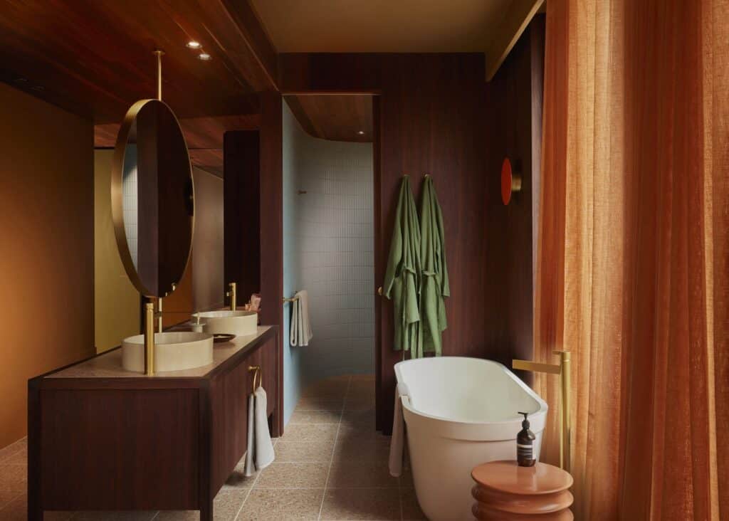 Tawny bathroom at Melbourne Place Hotel with a jarrah vanity, round brass-framed mirror, freestanding bath, green robes and a curved tiled shower