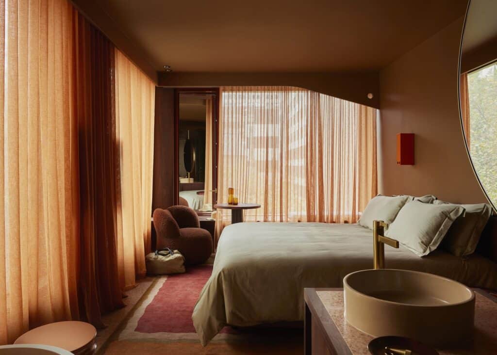 West-facing guest room at Melbourne Place Hotel glowing behind amber linen curtains, with a boucle armchair, pink rug and round basin